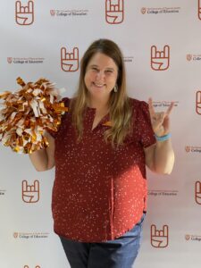 A smiling woman holds a bright silver and orange pom pom in her right hand and raises her left hand in the "hook em horns" symbol, with forefinger and pinkie fingers raised. 