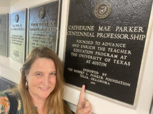 Selfie of Stephanie Cawthon leading up against a wall of plaques pointing her finger upward at the bronze plaque that commemorates her endowed professorship.