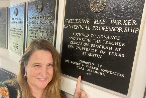 Selfie of Stephanie Cawthon leading up against a wall of plaques pointing her finger upward at the bronze plaque that commemorates her endowed professorship.