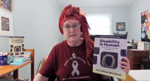 a middle-aged, cisgender, white female wearing glasses, a colorful headscarf, and t-shirt that reads "Rare and Resilient, Hidden Disability Awareness" with the rare disease zebra ribbon. She is holding up a copy of the Disability Is Human workbook.