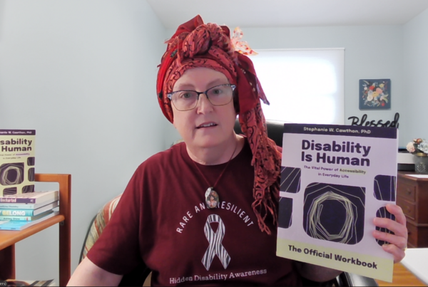 a middle-aged, cisgender, white female wearing glasses, a colorful headscarf, and t-shirt that reads "Rare and Resilient, Hidden Disability Awareness" with the rare disease zebra ribbon. She is holding up a copy of the Disability Is Human workbook.