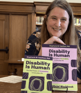 A close up of Stephanie behind her two books on display. She is in a library with lots of wooden paneling and the books are both Disability Is Human, the main book is one bright green and the Official Workbook is in lilac, both with dark purple font and decorative accents.