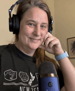 A tired looking close up of Stephanie with a headset on and in front of a blue mic. She has her hand up to her face and is wearing a grey shirt with white detail and a small smile.