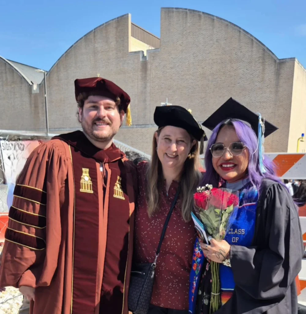 A man and two women are posing for a picture with graduate regalia and mortar boards in front of a brick building and a construction site. The are in the sunshine and have big smiles.