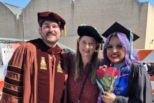 A man and two women are posing for a picture with graduate regalia and mortar boards in front of a brick building and a construction site. The are in the sunshine and have big smiles.