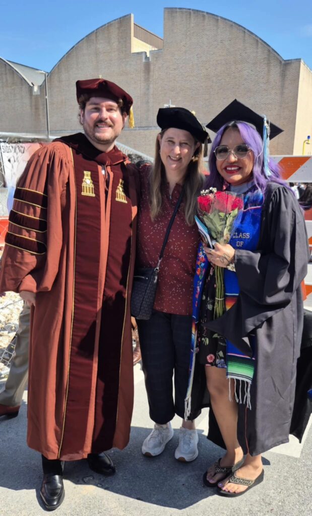 A man and two women are posing for a picture with graduate regalia and mortar boards in front of a brick building and a construction site. The are in the sunshine and have big smiles. 