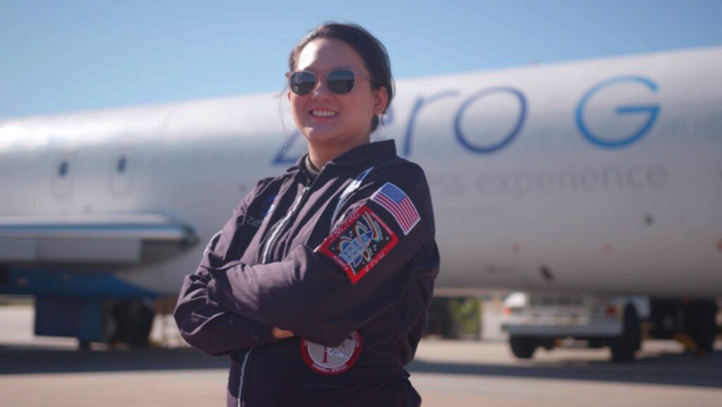 Asian woman in a flight suit with flags and patches wearing sunglasses and standing in a power pose in front of a zero gravity plane.