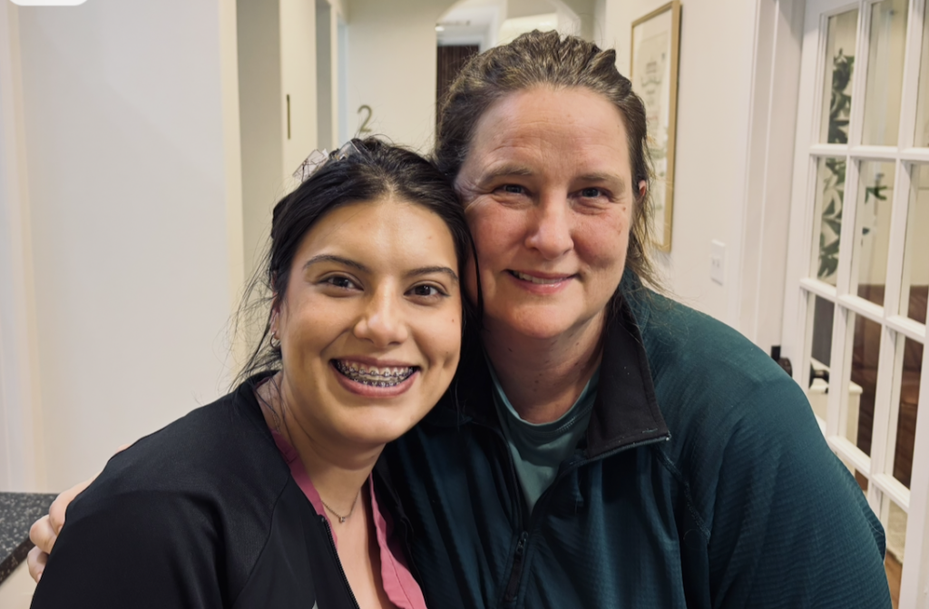 Two women smiling at the camera. The one on the left has braces and a huge grin and is wearing black and pink scrubs. Stephanie is on the right with small smile (she has a mouth full of novocaine) and is wearing a bright green pullover. Both have tousled hair.