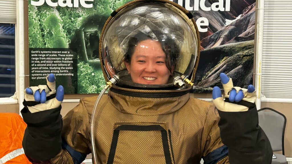 Asian female smiling in a space suit with a clear helmet. She has her hands up in an “I love you” sign from American Sign Language.