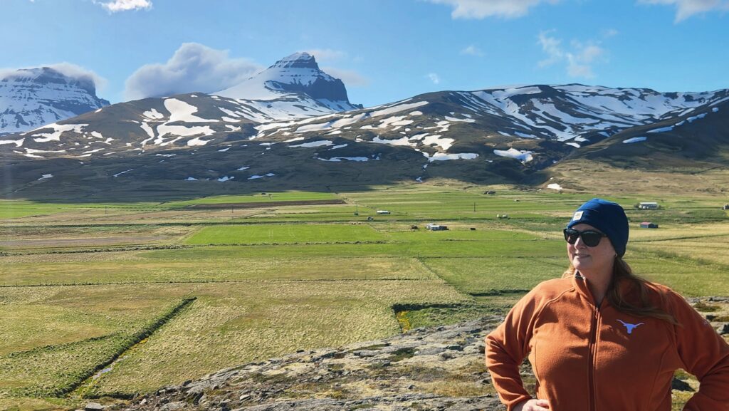 Stephanie looking off in the distance wearing an UT Branded orange fleece, She is standing on rocky outcropping with mountains and fields in the distance. It is a glorious vista.