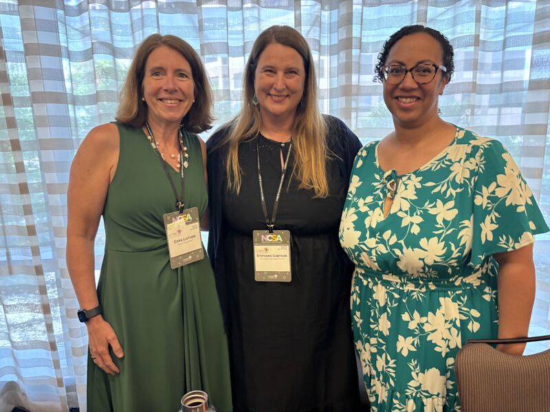 Three women in a pose in front of hotel conference room window, all smiling and wearing business conference dresses in a range of colors and patterns. Two are wearing conference badges that say NCSA 2026.